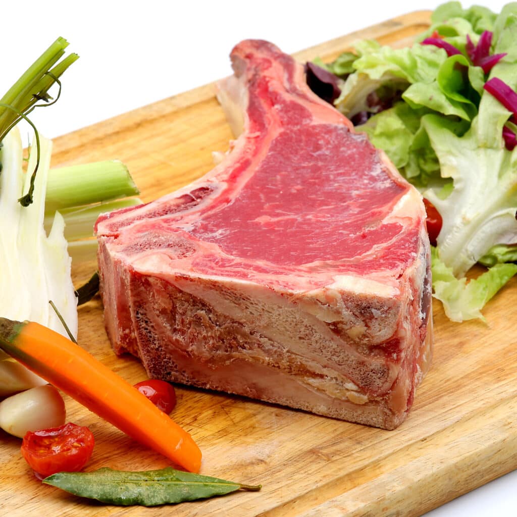 Épaisse côte de bœuf crue sur une planche à découper en bois, entourée de légumes frais : fenouil, céleri, ail, tomate et salade.
