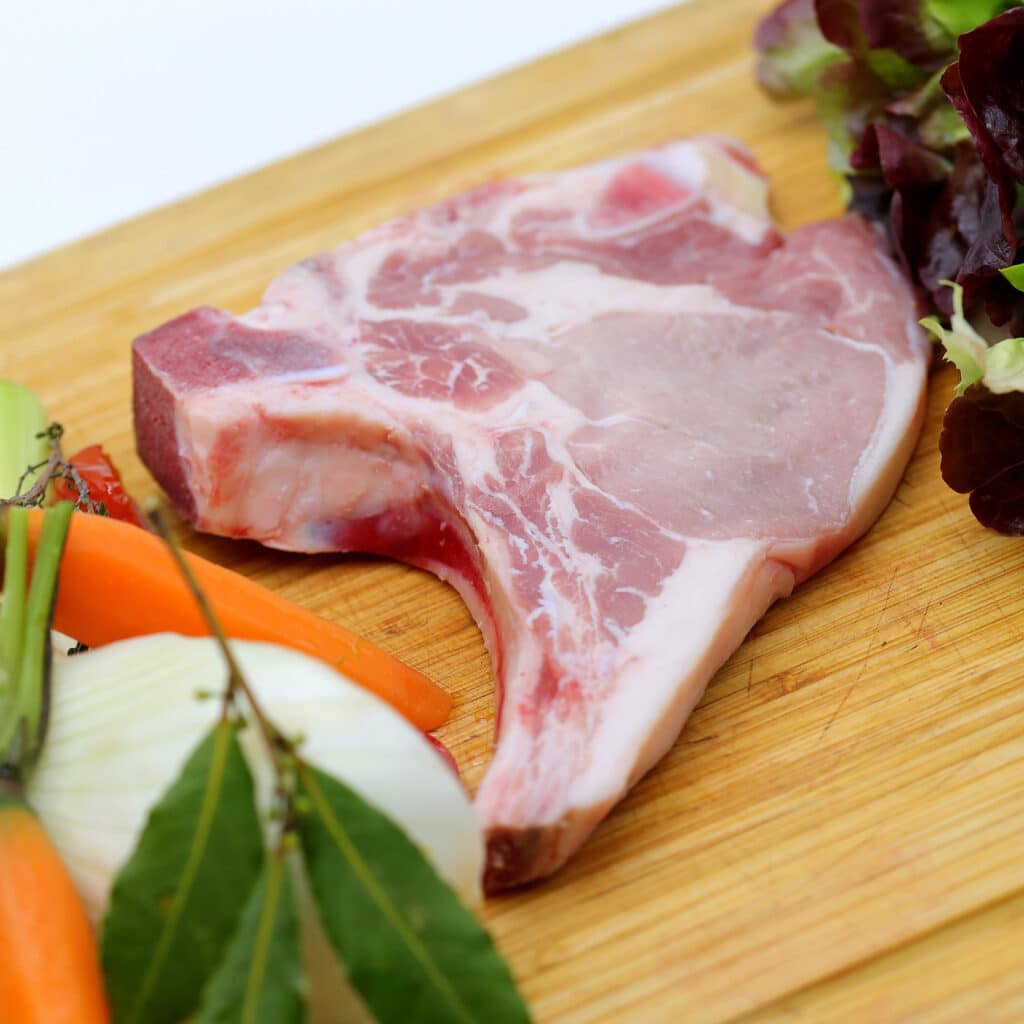 Côte de porc fraîche et légumes de saison sur planche Côte de porc crue sur os, marbrée, sur une planche à découper en bois, avec carottes, oignon, laurier et salade pour la préparation.