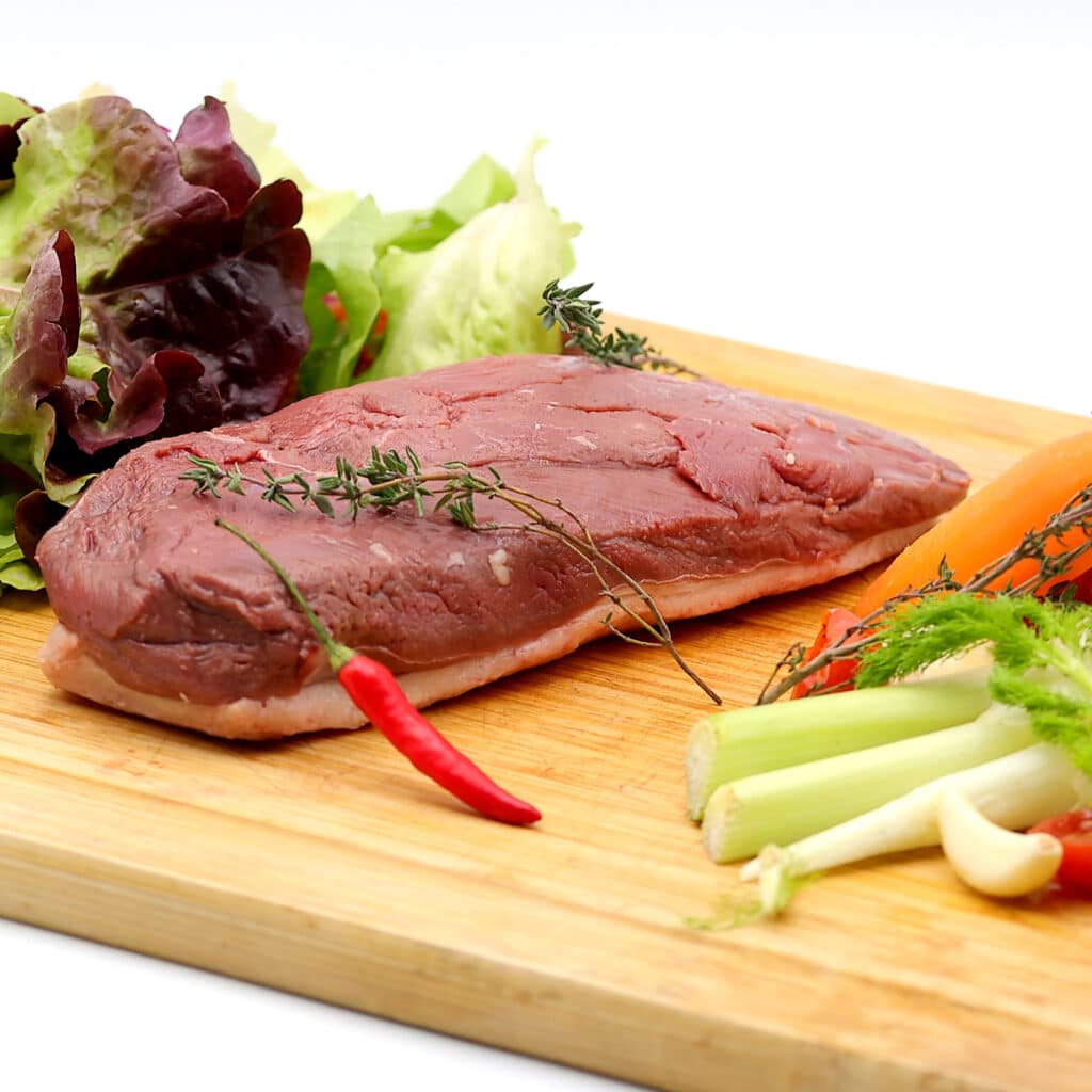 Filet de canard cru sur planche en bois, avec du thym, un piment rouge, des légumes frais (céleri, carotte, ail et salade).