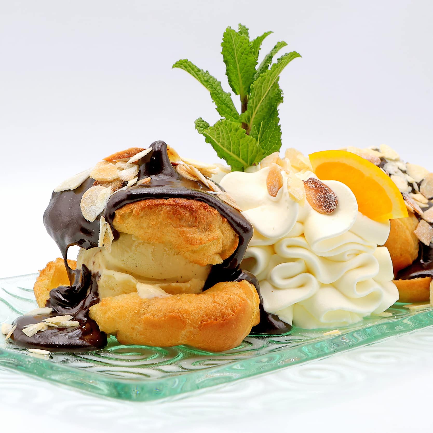 Profiteroles à la glace vanille et sauce chocolat. Garniture de crème fouettée, amandes, menthe fraîche et orange sur une assiette en verre.