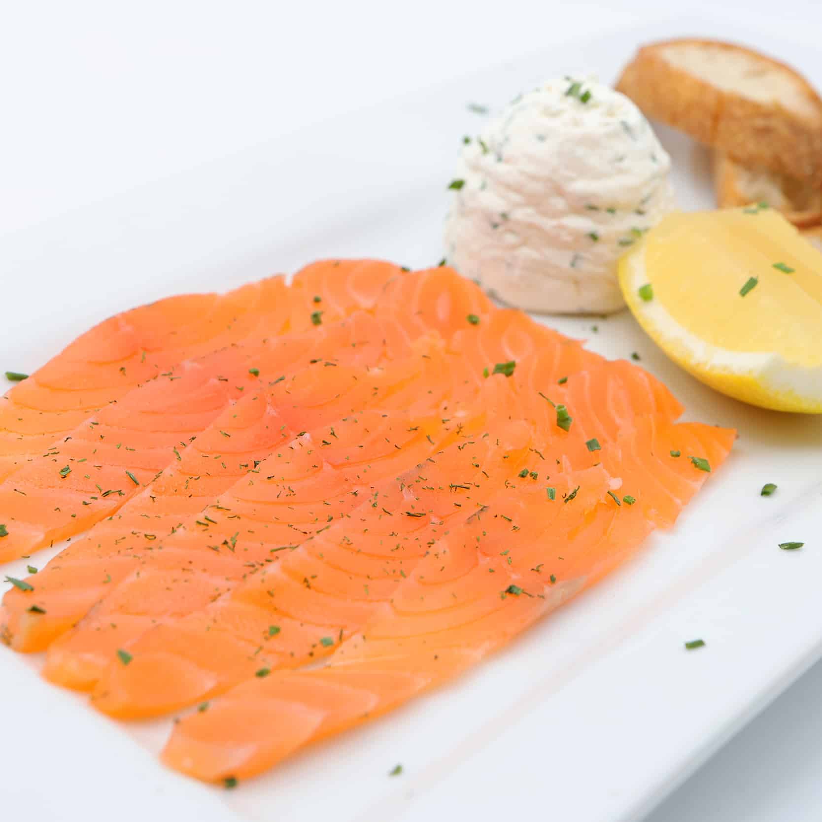 Tranches de saumon fumé à l'aneth, accompagnées de fromage frais à la ciboulette, d'un quartier de citron et de pain grillé sur assiette blanche.