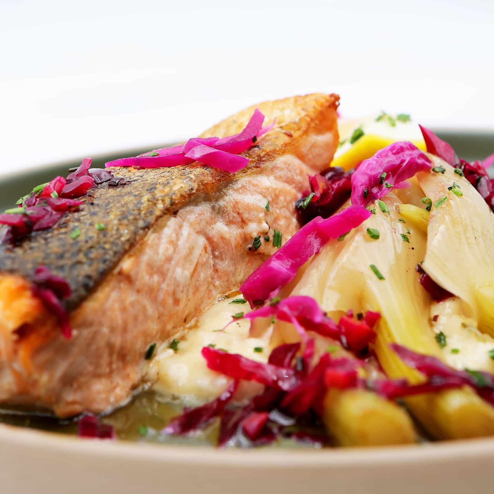 Saumon à la peau croustillante, fenouil braisé et chou mariné. Filet de saumon à peau croustillante, servi sur une purée crémeuse avec des morceaux de fenouil braisé, et garni de chou rouge mariné vif.